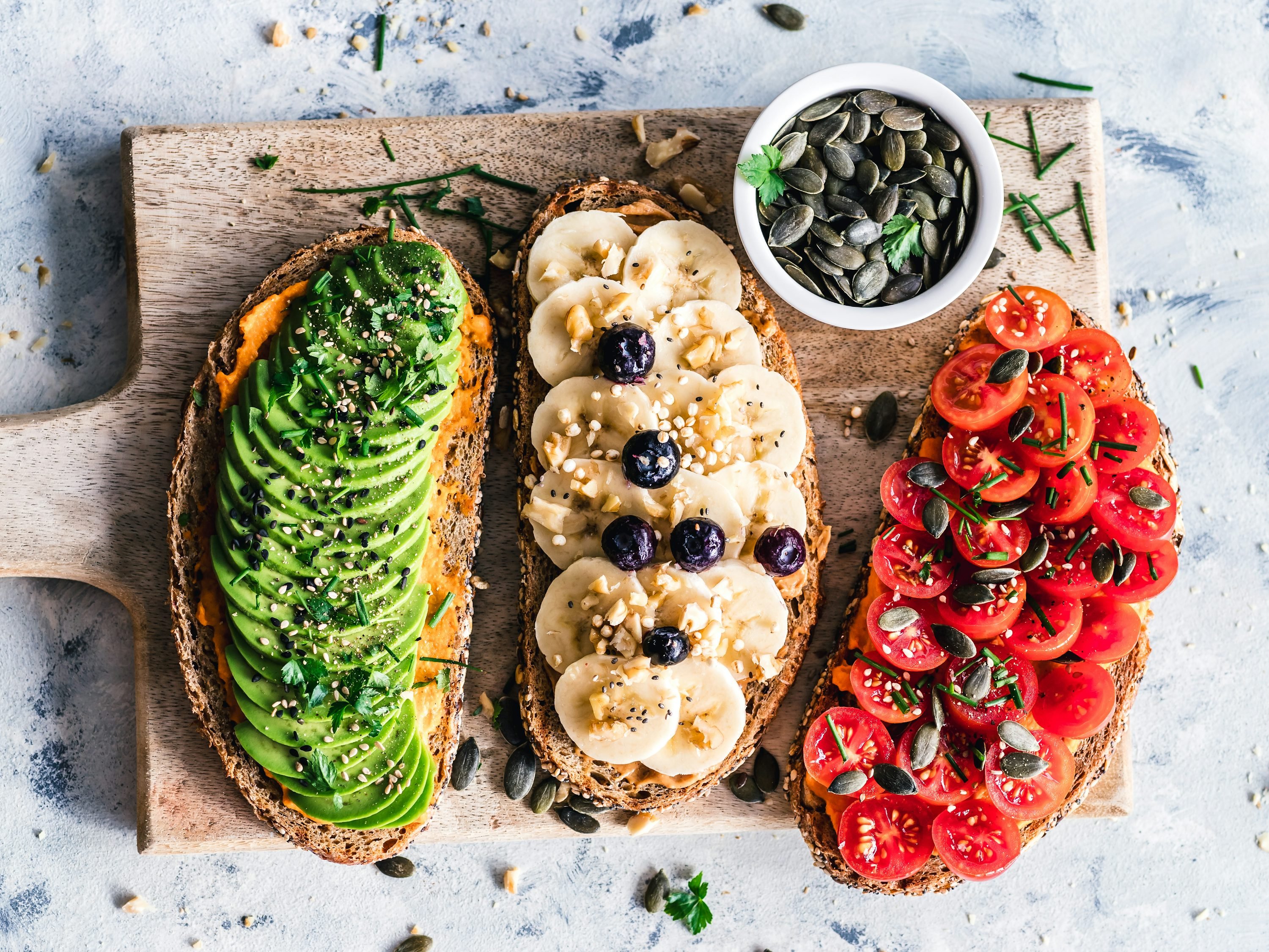 En la imagen se ven tres mitades de pan tostado con toppings: aguacate en la izquierda, plátano y arándanos en la mitad central